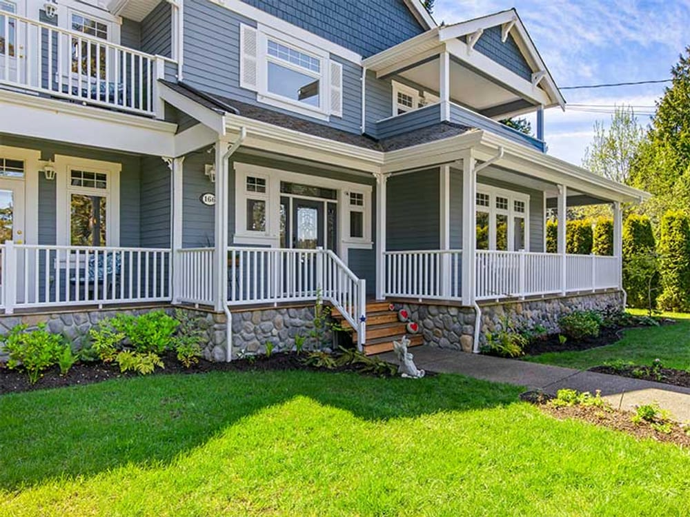 A blue-grey shingled house with white trim and a wrap-around veranda, seen across its lawn.