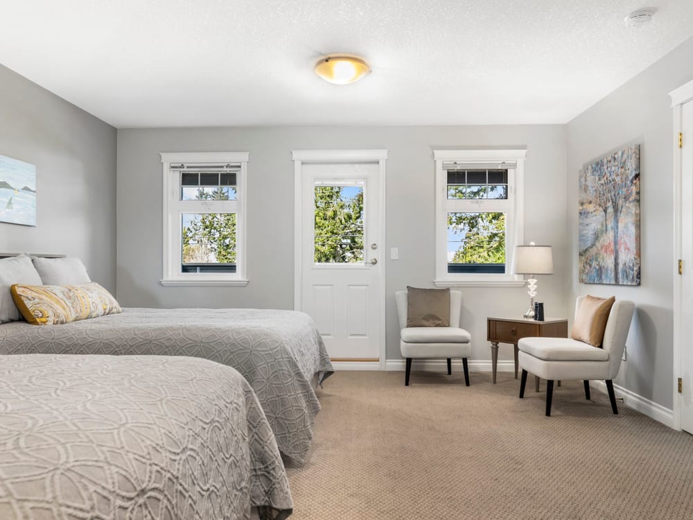 A grey and white guest room with two queen beds, a private door between two windows, and a pair of cream armchairs beside a lamp.