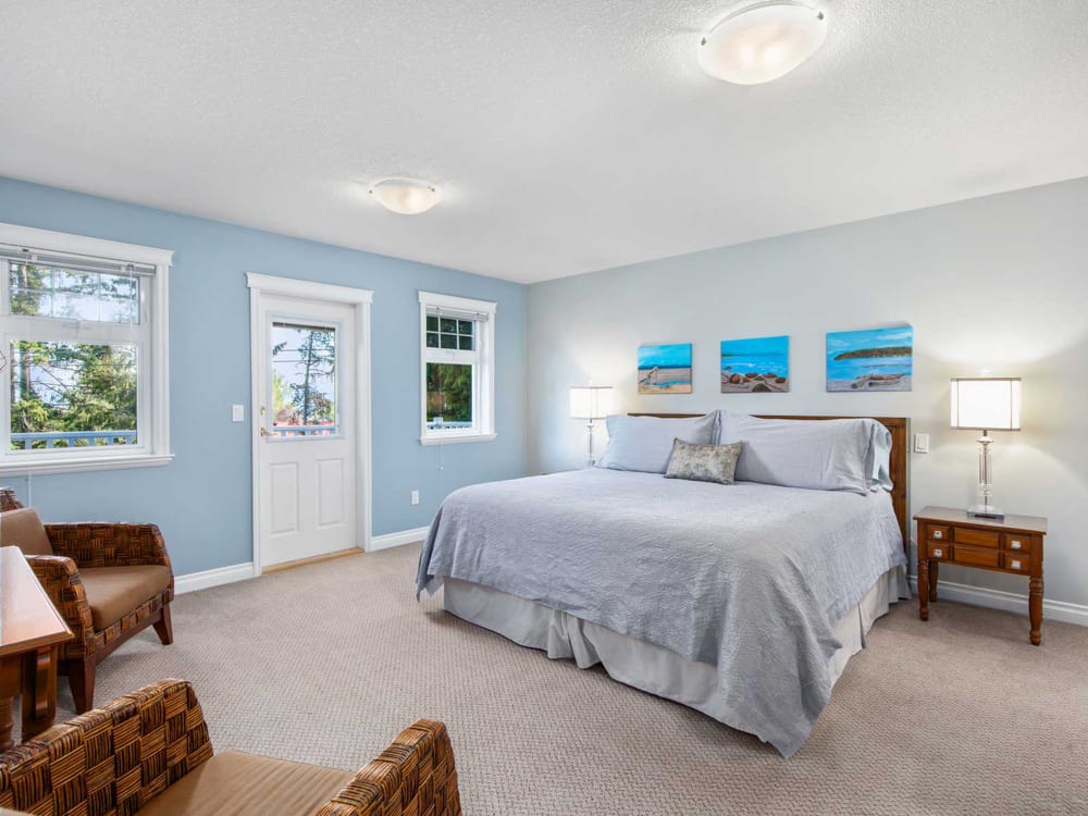 A pale blue guest room with a king bed under three small seascape canvases, woven armchairs and a door to the balcony.