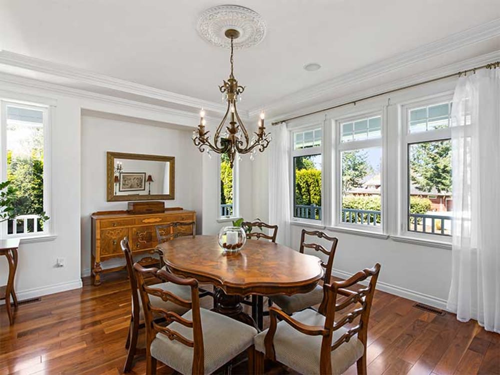 A dining room with an oval wooden table under a chandelier, windows along two walls and a sideboard against the third.