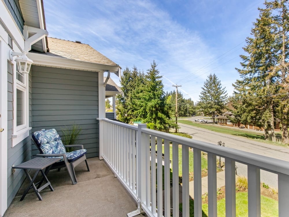 The veranda along the side of the house, a cushioned chair beside the white railing and the road and firs beyond.