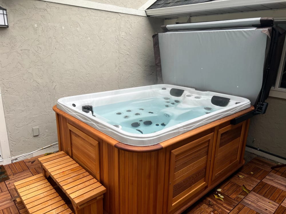 A cedar-set salt-water hot tub on a brick patio beside the house, its cover folded back.