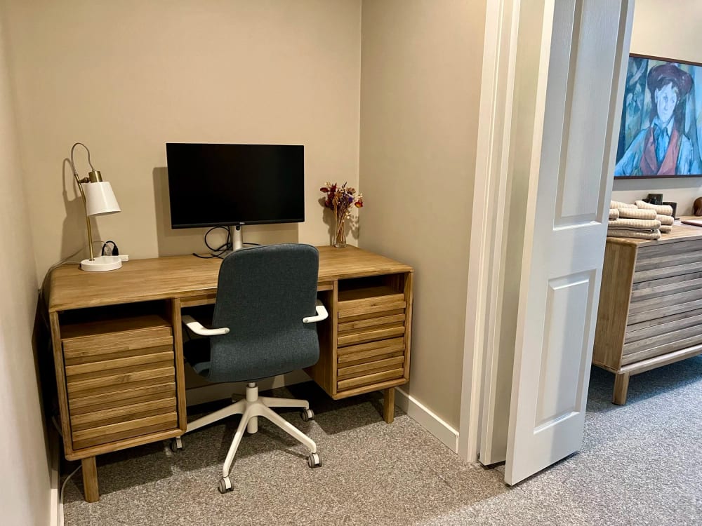 A wooden desk with a monitor and a lamp beside the doorway through to the bedroom.