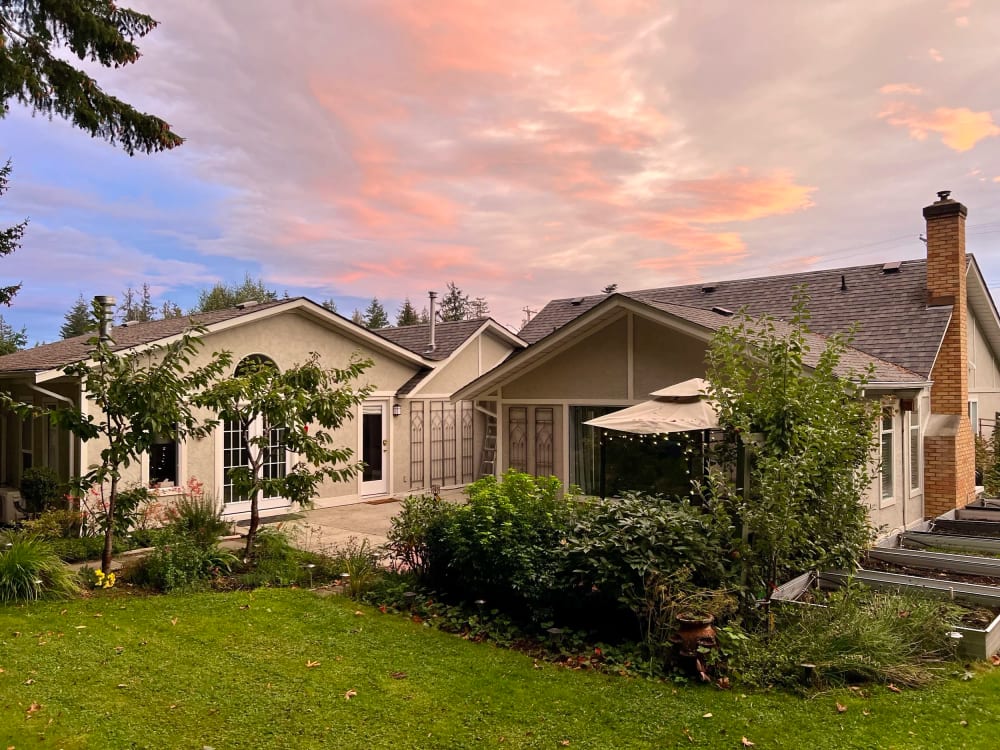 A single-storey house with a peaked roof above a lawn, the garden borders lit by the last of the sun.