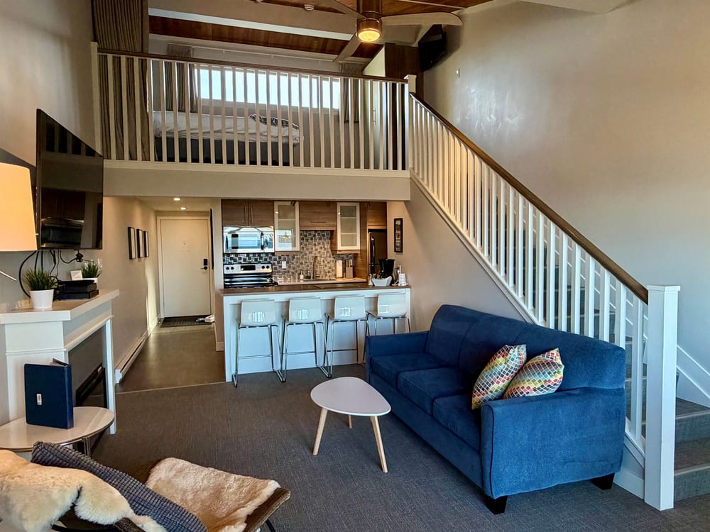 The open living space of the loft seen from the door, with the loft floor overhanging it and windows to the water.