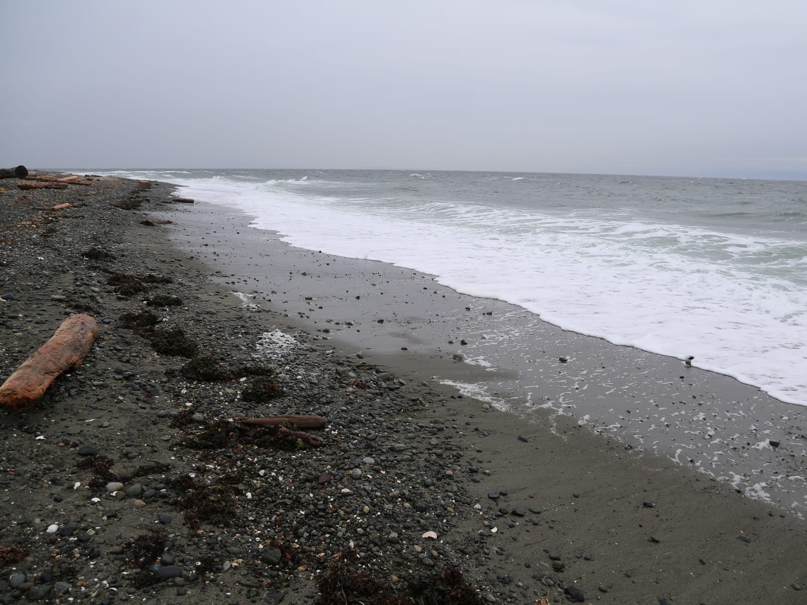 A wide cobble-and-sand beach under a soft sky, the tide far out and the mountains faint across the strait.