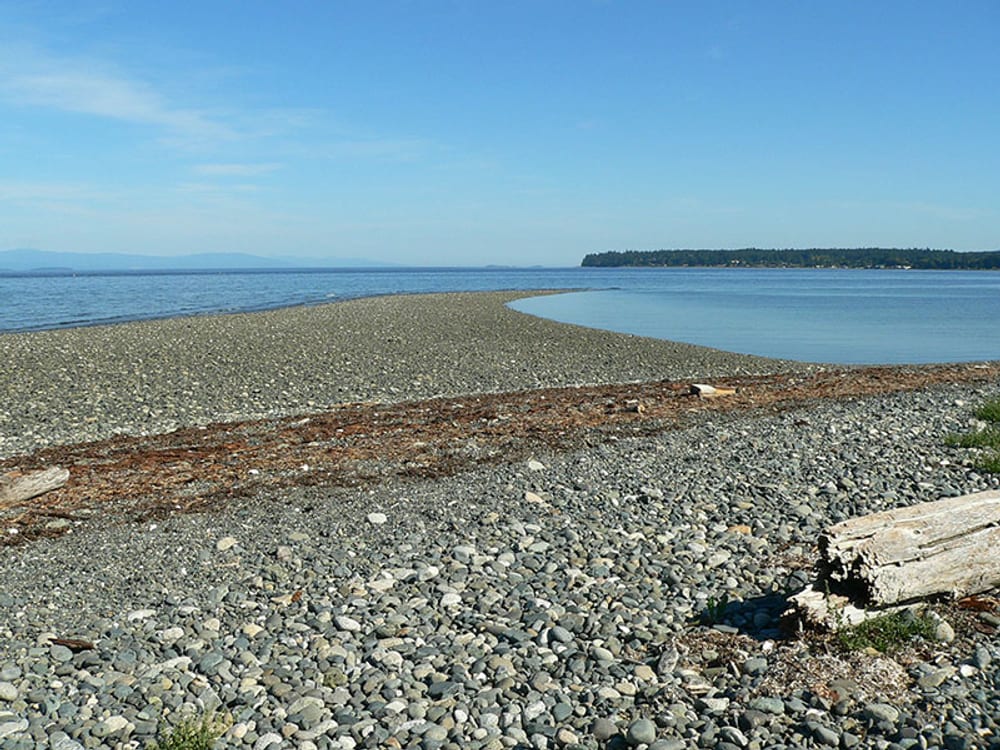A long gravel spit reaching into the bay at low tide, the far shore beyond.